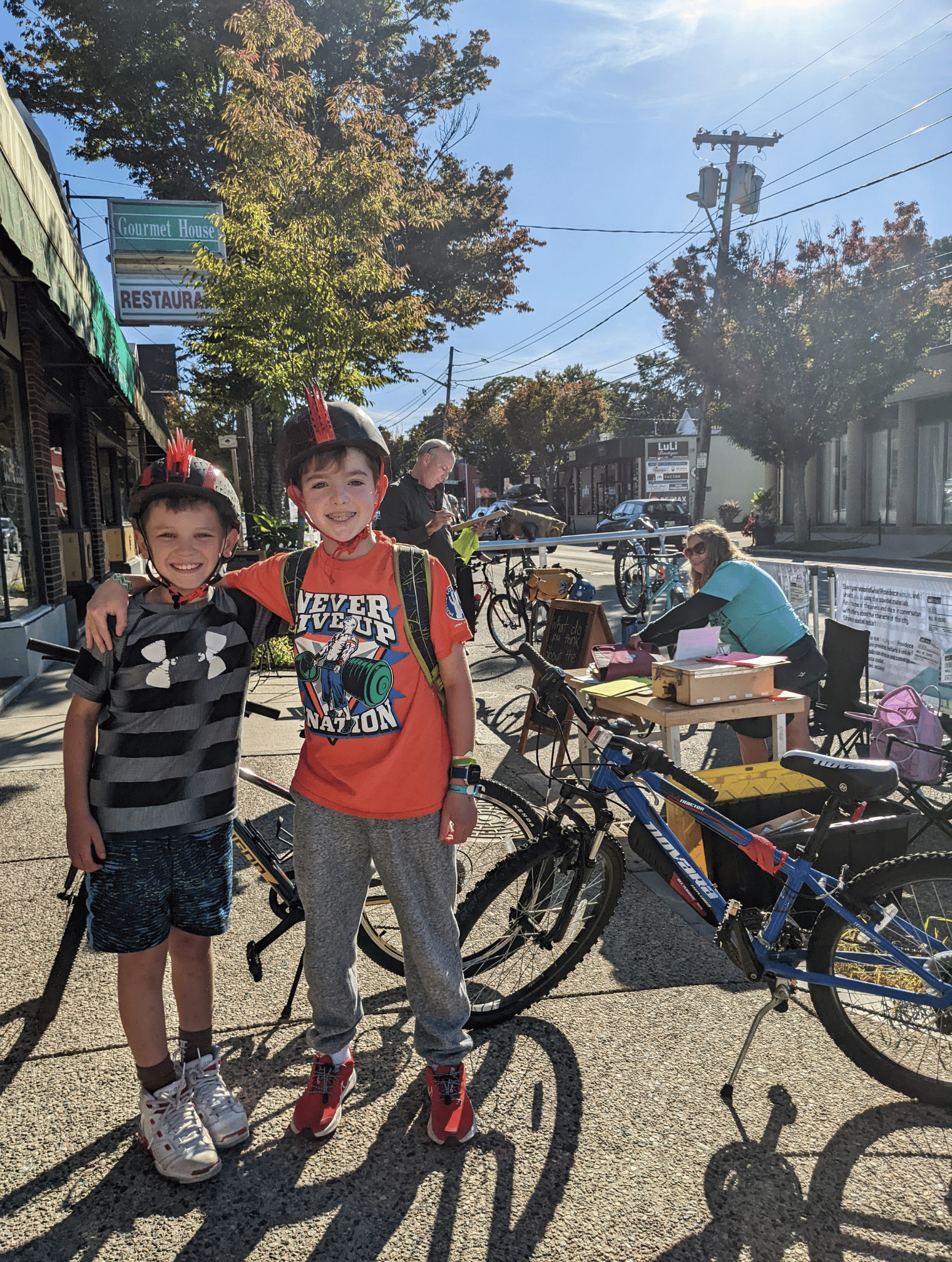 Two boys stand in front of their bikes adjacent to the Hope Street temporary trail with their arms around each other. One is wearing shorts and a gray and black t-shirt, the other is wearing grey pants and an orange graphic t-shirt. Both are wearing black helmets with bright red plastic mohawks emerging from the top.