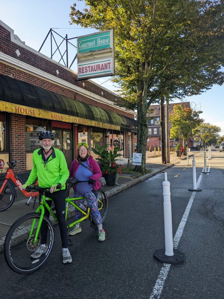 Two adults straddling a bright green tandem bicycle stand in the temporary trail in front of a restaurant. In the front is a man with a green high-vis shirt and black pants. In the rear is a woman with purple pants and shirt and a maroon sweater.