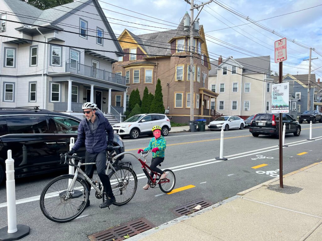 View of the temporary trail on a bright sunny day with three-story mutilfamily houses in the background, and the temporary trail in the foreground lined with white bollards spaced regularly and bright yellow centerline stripes. A man in a blue jacket on a gray bicycle, accompanied by a child atop a bike trailer wearing a bright aqua jacket, red gloves and a pink gaiter, travels in the bike lane.