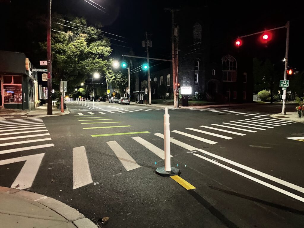 Nighttime view of an intersection with green and red traffic lights facing opposite directions. Bright white and green stripes on the black pavement designate crossing for pedestrians and bicycles. White bollards stretch into the distance designating the temporary trail.