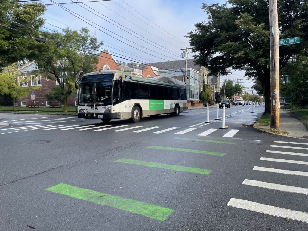View of a tree lined street under sunny skies. Green stripes painted on the pavement parallel the white stripes of crosswalks as the temporary trail in the background reaches an intersection. A transit bus drives in the travel lane.