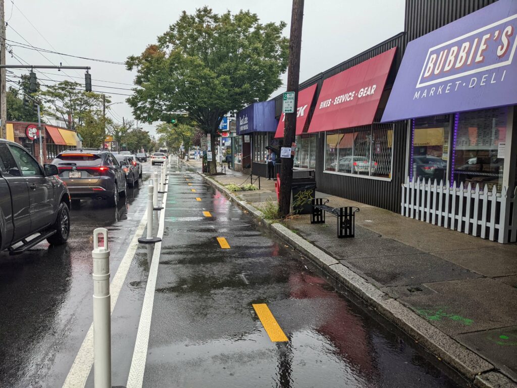 View of the tree-lined temporary trail, awash in recent rainfall under still cloudy skies, with bright yellow centerline markers, white boundary markers and white bollards spaced evenly. Colorful awnings of small local businesses line the sidewalk.